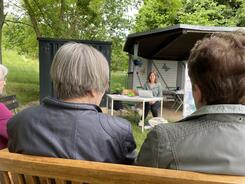 Drei Teilnehmerinnen sitzen drau&szlig;en in einem Garten auf B&auml;nken und schauen zu Theresa, die an einem Tisch mit Laptop sitzt.