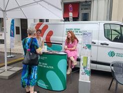 Eine Dame steht vor dem Infomobil und blickt zu den beiden Referentinnen Theresa und Lena, die auf Barhockern sitzen.