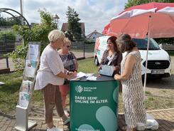 Theresa zeigt drei Besucherinnen am Infostand etwas auf dem Tablet.