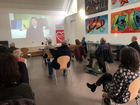 Personen sitzen mit Abstand auf St&uuml;hlen in einem Raum und schauen Video auf Leinwand