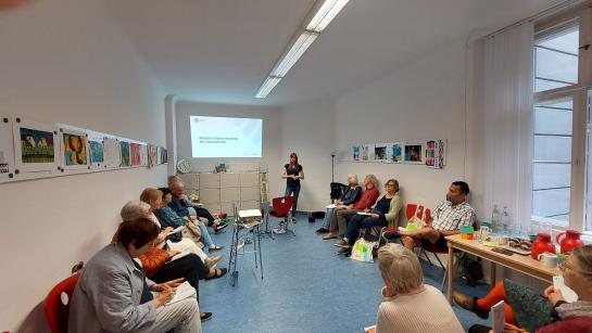 Blick in einem Raum, in dem rund 14 St&uuml;hle mit Senior:innen besetzt sind. Sie lauschen einer Vortragenden. 