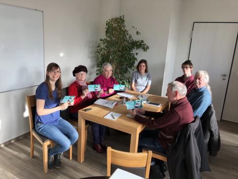 Die Referentinnen Petra und Theresa sitzen gemeinsam mit f&uuml;nf Teilnehmenden der Veranstaltung an einem Tisch und halten Flyer vom Digitalen Engel. Einige l&auml;cheln in die Kamera. 