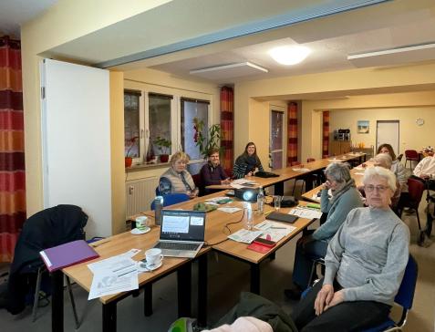 Sechs Senior:innen, Petra und Theresa sitzen an Tischen und l&auml;cheln in die Kamera.