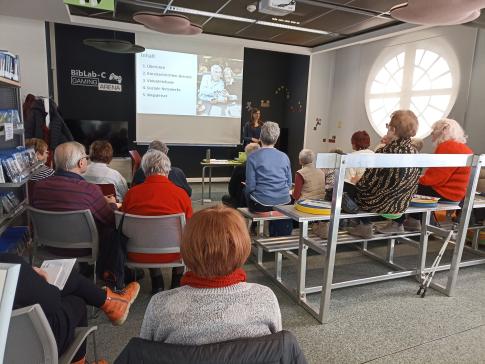 Senior:innen sitzen auf St&uuml;hlen und B&auml;nken (von hinten fotografiert). Der Blick richtet sich nach vorne auf die Referentin vom Digitalen Engel mit einer Pr&auml;sentation.
