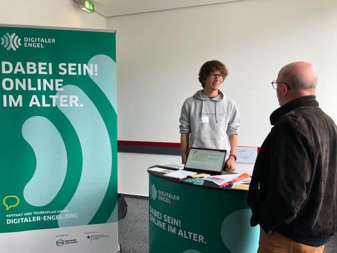 Mobilreferent Johannes ist im Gespr&auml;ch mit einem Fachbesucher am Messestand des Digitalen Engel. Auf der linken Seite steht ein Roll Up mit der Aufschrift "Dabei sein! Online im Alter."