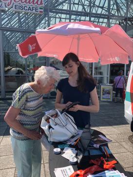 Petra zeigt einer Seniorin drau&szlig;en am Infostand Dinge am Smartphone.