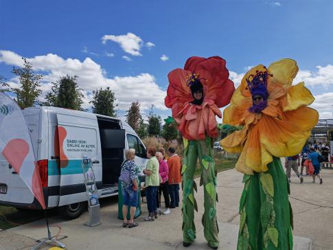 Zwei als &uuml;bergro&szlig;e Blumen verkleidete Maskottchen des Landesseniorentages stehen neben unserem Infomobil an dem sich einige Besucherinnen mit Johanna unterhalten 