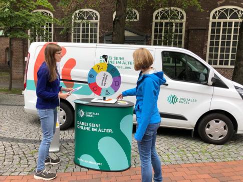 Petra steht mit Melanie Sing von der Stadt Elmshorn am Gl&uuml;cksrad vor dem Info-Mobil. Frau Sing dreht das Gl&uuml;cksrad und Petra h&auml;lt Zettel mit Fragen in der Hand.