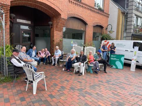 Das Info-Mobil steht vor dem Caf&eacute;. Petra bespricht mit einer Seniorin ein individuelles Thema. Daneben sitzen viele Senior:innen auf St&uuml;hlen und trinken Kaffee.