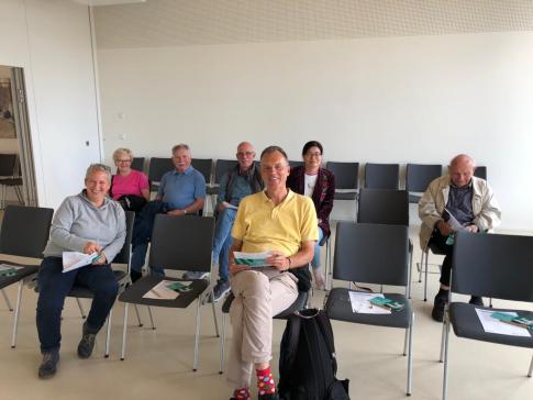 Sieben Personen sitzen auf St&uuml;hlen und gucken in die Kamera.