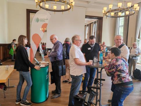 Petra steht am Info-Stand des Digitalen Engel w&auml;hrend der Markt der M&ouml;glichkeiten und ist im Gespr&auml;ch mit einem &auml;lteren Herrn. Neben dem Stand stehen vier &auml;ltere Menschen um einen runden Tisch und unterhalten sich. 