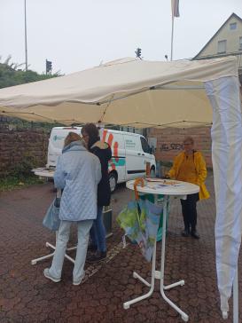 Petra steht mit einer Dame am Info-Stand. Der Info-Stand ist mit einem Pavillon vor dem Regen gesch&uuml;tzt.