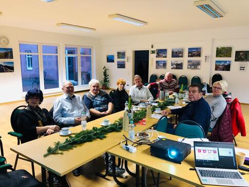 Gruppenbild der Teilnehmenden. Acht Damen und Herren sitzen an einem winterlich geschm&uuml;cktem Tisch. Im Vordergrund sind ein Laptop und ein Beamer zu sehen.