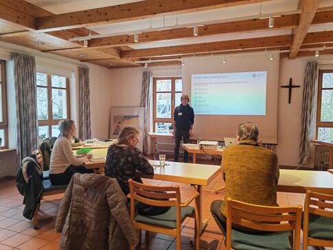 Die Teilnehmenden sitzen im Raum und blicken zur Pr&auml;sentation an der Leinwand und zu unserem Referenten Johannes der neben der Leinwand steht