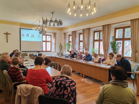 Die Teilnehmenden sitzen an einem gro&szlig;en Tisch im Sitzungssaal und schreiben