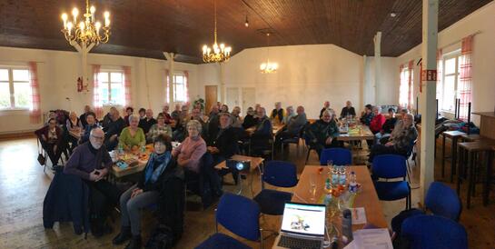 Ein Panorama der knapp 50 Teilnehmenden, die in einem gro&szlig;en Raum an verschiedenen Gruppentischen sitzen. Im Vordergrund steht ein Laptop.