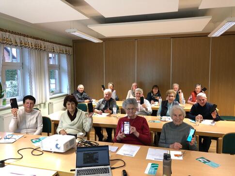Zw&ouml;lf Seniorinnen und zwei Senioren sitzen an Tischen und halten ihr eigenes Smartphone in die Kamera.