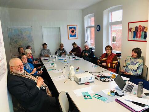 Elf Senior:innen sitzen an einem gro&szlig;en Tisch im Viereck. Einige gucken in die Kamera. Andere halten ein Smartphine in der Hand. Auf dem  Tisch stehen ein Beamer und ein Laptop von Petra.