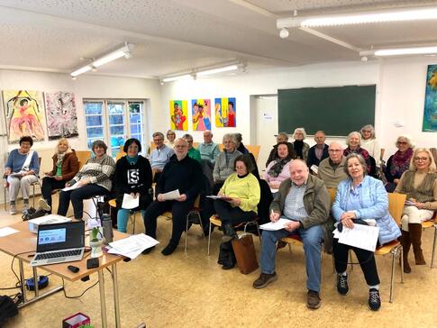 24 der knapp &uuml;ber 30 Teilnehmenden sind auf dem Bild zu sehen. Sie sitzen auf St&uuml;hlen und gucken in die Kamera nach vorne. Einige halten Unterlagen in den H&auml;nden. Im vorderen Bildabschnitt steht ein Laptop und ein Beamer.