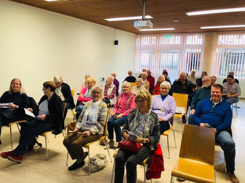 Knapp 30 Senior:innen sitzen auf St&uuml;hlen in f&uuml;nf Reihen ohne Tische. Einige gucken in die Kamera - andere gucken weg.