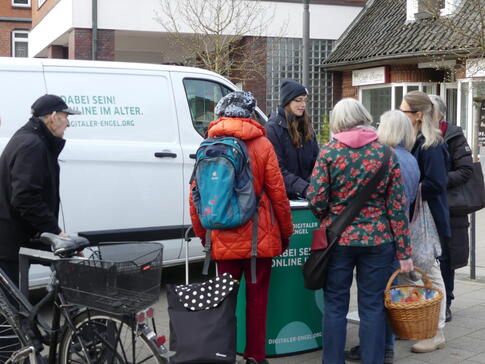 Pera steht in Winterjacke und M&uuml;tze vor dem Info-Mobil am Info-Stand. Um den Stand herum stehen f&uuml;nf Damen mit Einkaufstaschen und ein Herr mit einem Fahrrad.