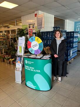 Theresa steht am Infomobilstand im Combi Supermarkt. Links hinter ihr sind Blumen, direkt hinter ihr Wassergetr&auml;nkekisten.