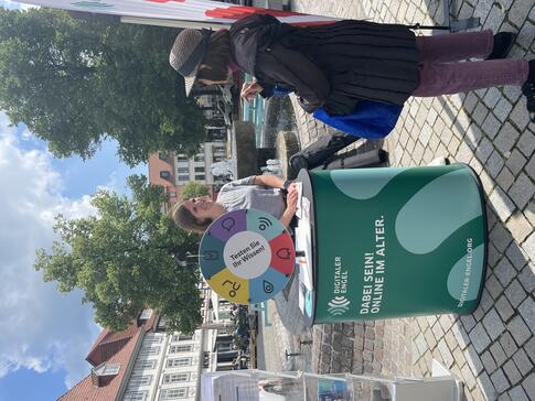 Theresa und eine Besucherin unterhalten sich am Infostand. Hinter ihnen sind ein Brunnen sowie H&auml;user und zwei B&auml;ume zu sehen.