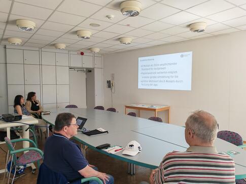 Die Teilnehmenden sitzen an einem gro&szlig;en Tisch im Raum und blicken zur Pr&auml;sentation an der Wand