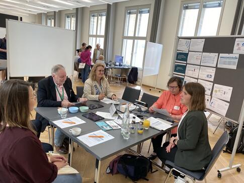 Theresa und die Besuchenden sitzen um einen viereckigen Tisch. Theresa erkl&auml;rt Ihnen Digitale Gesundheitsanwendungen.