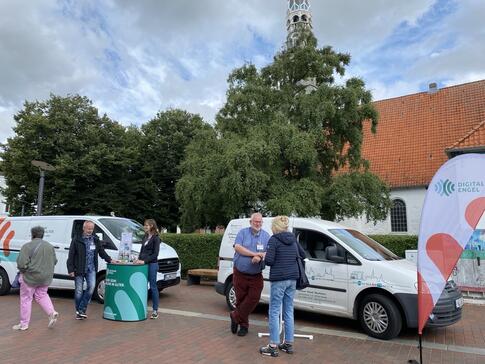 Das Info-Mobil (links) steht neben dem Mobil vom offenen Kanal Westk&uuml;ste (rechts). Vor dem rechten Mobil steht Andreas mit einer Dame. Links stehen Horst und Petra am Info-Stand. Eine Seniorin verl&auml;sst gerade den Stand.