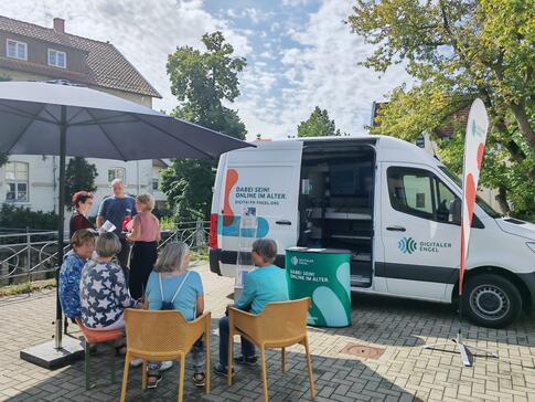 Mehrere Besucherinnen sitzen auf St&uuml;hlen vor dem Infomobil und unterhalten sich, au&szlig;erdem steht Frau R&ouml;the mit einer weiteren Besucherin und einem Besucher an einem Stehtisch