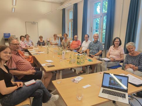 Die Teilnehmenden der Veranstaltung sitzen an einem gro&szlig;en zusammengeschobenen Tisch und blicken nach vorne