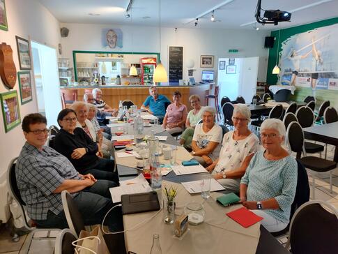 Zw&ouml;lf Teilnehmende (neun Frauen und drei M&auml;nner) sitzen an einem langem Tisch im Vereinsheim und gucken nach vorne in die Kamera.