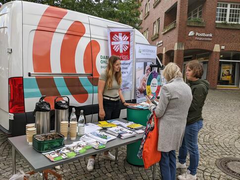 Theresa und eine Besucherin, die links neben ihr steht, sind von hinten zu sehen und h&ouml;ren einer Caritas-Mitarbeiterin zu, die am gemeinsamen Stand vor dem Infomobil steht.