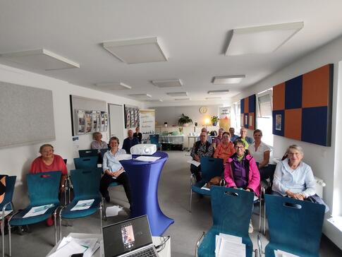 Auf der linken und rechte Seite des Veranstaltungsraumen sitzen elf Teilnehmende sowie die Ansprechpartnerinnen des Senioren- und Pflegest&uuml;tzpunkt. In der Mitte des Raumes steht ein Beamer und ganz vorne im Bild ist ein Laptop zu sehen. 