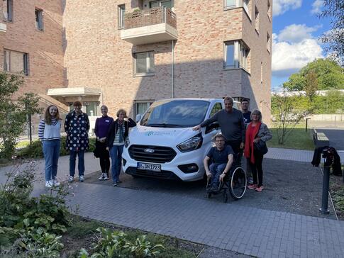 4 Teilnehmerinnen gemischten Alters stehen links neben dem Infomobil sowie drei Teilnehmer und eine Teilnehmerin rechts neben dem Infomobil.  Das Mobil steht auf einer sandigen Fl&auml;che umgeben von einem gepflasterten Weg. Links im Hintergrund ist ein mehrgesch&ouml;ssiges, neueres Haus, rechts im Hintergrund  B&auml;ume, Gras und Wolken. 