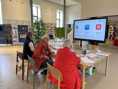 Die drei Teilnehmerinnen sitzen an einem gro&szlig;en Tisch und sind von hinten zu sehen. Die Veranstaltung ist in einem gr&ouml;&szlig;eren B&uuml;chereisaal. Vorne ist die Pr&auml;sentationsfolie zum Thema App herunterladen auf einem Smartboard abgebildet.
