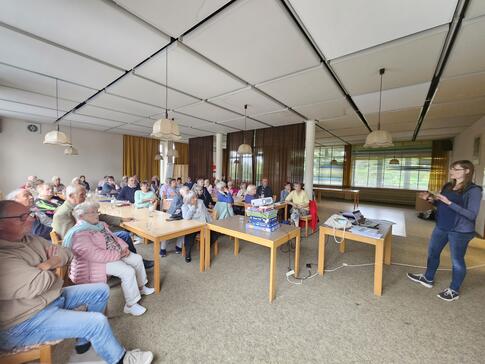 Auf der linken Seite des Bildes sitzen 45 Seniorinnen und Senioren auf St&uuml;hlen an  Tischen. In der Mitte des Bildes steht ein Beamer auf Spiele-Packungen. Rechts im Bild steht Petra mit ihrem Laptop und erkl&auml;rt etwas. 