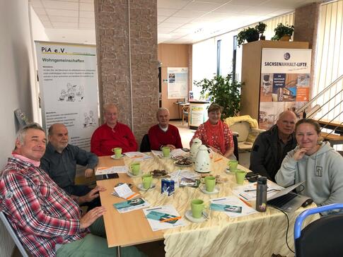 Sechs Senior:innen (f&uuml;nf M&auml;nner und eine Frau) und eine Ansprechpartnerin von PiA sitzen an einem Tisch im Kreis und gucken nach vorne in die Kamera.