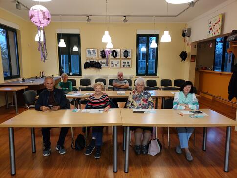 Im Veranstaltungsraum stehen Tische in zwei Reihen. In der ersten Reihe sitzen vier Seniorinnen und ein Senior (links). In der zweiten Reihe sitzt eine Seniorin (links) und ein Senior (miittig). Sie blicken alle nach vorne in die Kamera. Einige l&auml;cheln.