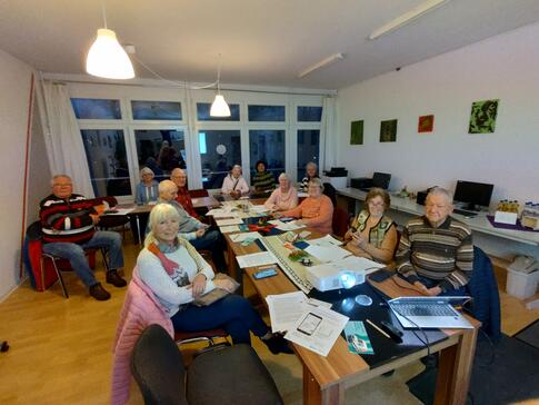 In einem Raum im MGH Buntes Haus sitzen vier &auml;ltere M&auml;nner und acht &auml;ltere Frauen an einem l&auml;nglichen Tisch. Die meisten blicken nach vorne in die Kamera. Auf dem Tisch im vorderen Bildrand stehen ein Beamer und ein Laptop.