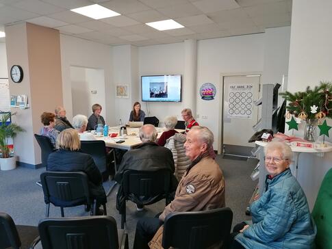 Im ROWI Stadtb&uuml;ro sitzen insgesamt elf Personen. Im Hintergrund sitzen an einem Tisch Petra mit f&uuml;nf &auml;lteren Menschen. Im Vordergrund sitzen auf St&uuml;hlen vier weitere Senior:innen. Die beiden Senior:innen ganz vorne drehen sich um und blicken in die Kamera.