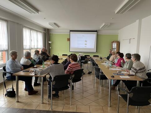 Die Teilnehmenden sitzen an zwei l&auml;ngeren Gruppentischen und schauen auf die Leinwand, auf der die Stationen des Digitalen Engels angezeigt werden. Die Wand hinter der Leinwand ist hellgr&uuml;n.