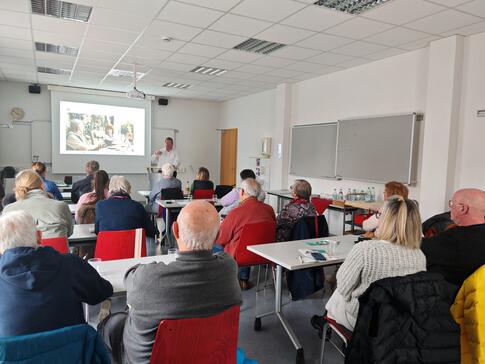 Mehrere Personen sitzen an Tischen im Raum und blicken nach Vorne zur Pr&auml;sentation auf einer Leinwand