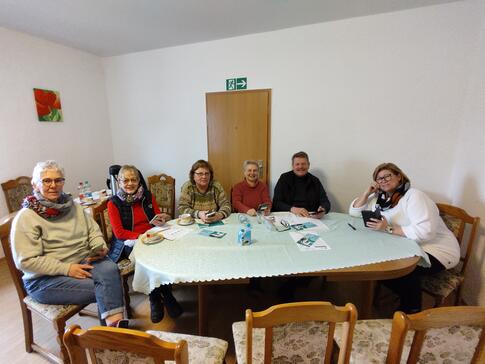 F&uuml;nf Seniorinnen und ein Senior sitzen an einem ovalen Tisch und l&auml;cheln vorne in die Kamera.