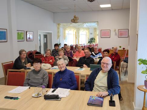 Im Plauderst&uuml;bchen sitzen in vier Reihen &uuml;ber 15 Senior:innen und blicken nach vorne. Einige l&auml;cheln.