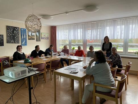 Die 8 Teilnehmenden sitzen an Tischen in einer U-Form. Theresa steht hinter ihnen und l&auml;chelt in die Kamera.