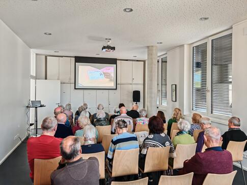 Mehrere Personen sitzen auf St&uuml;hlen im Raum und blicken nach Vorne auf eine Leinwand auf der ein Video abgespielt wird.