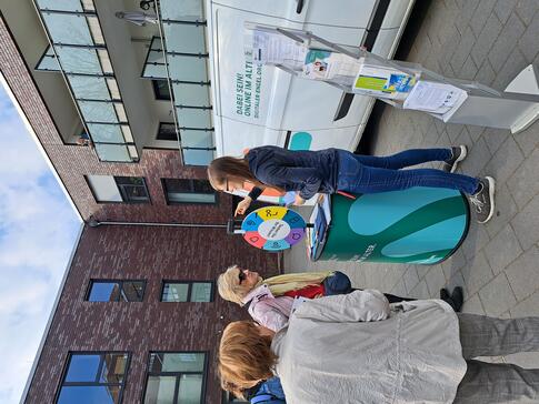 Petra steht mit dem Info-Stand vor dem Info-Mobil am Gl&uuml;cksrad. Drei Seniorinnen stehen um sie herum und eine Seniorin beantwortet eine Frage.
