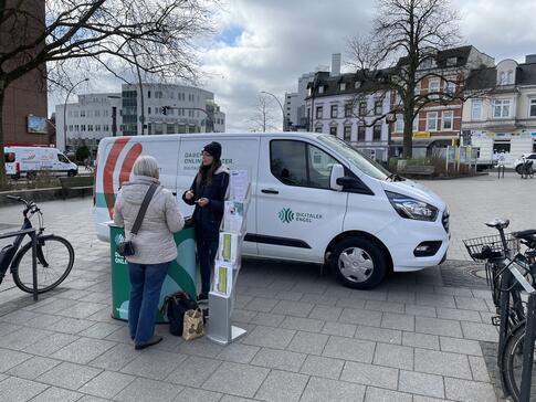 Petra steht mit einer Seniorin am Info-Stand vor dem Info-Mobil und ist mit ihr im Gespr&auml;ch. Am linken und rechten Bildrand steht jeweils ein Fahrrad.
